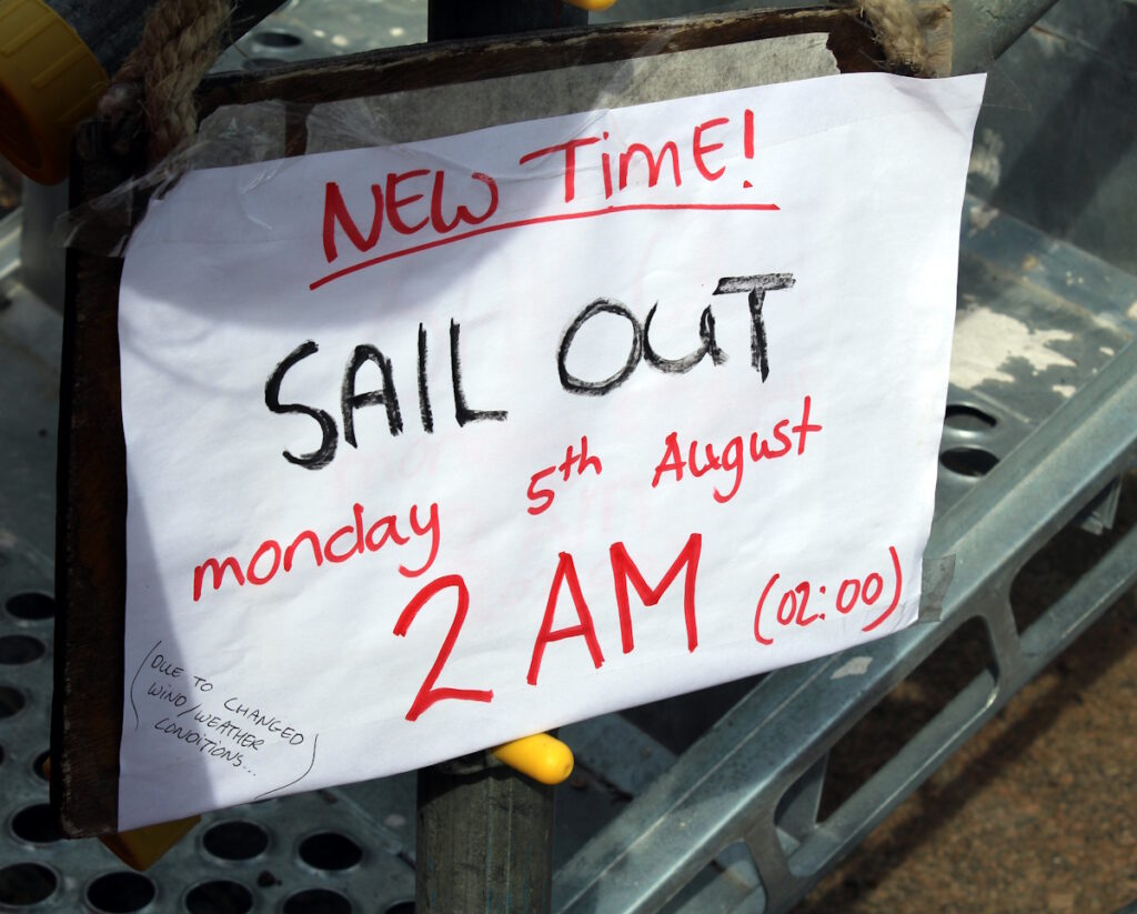 A sign on the Nao Victoria giving notice of its leaving time. The sign is handwritten in red and black ink on white paper. The sign reads 'New time! Sail out Monday 5th August 2AM (02:00) (due to changed wind/weather conditions).'