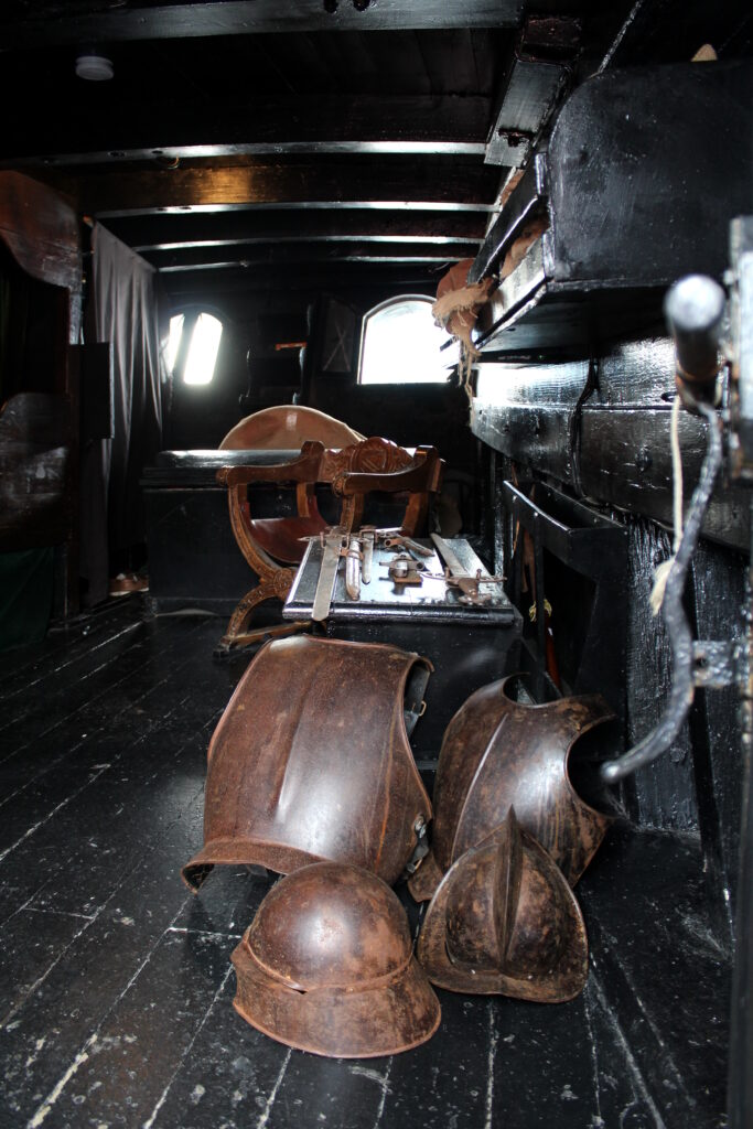 interior of the captain's cabin on the Nao Victoria. A desk and chair is visible. There are two armour body plates and two helmets on the floor of the cabin.