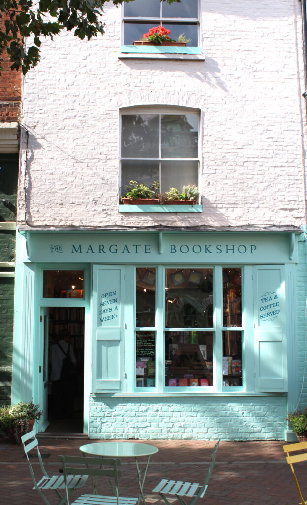 An image of the exterior of the Margate Bookshop in Margate. The storefront is turquoise blue and there are turquoise blue cafe tables and chairs in front of the shop.