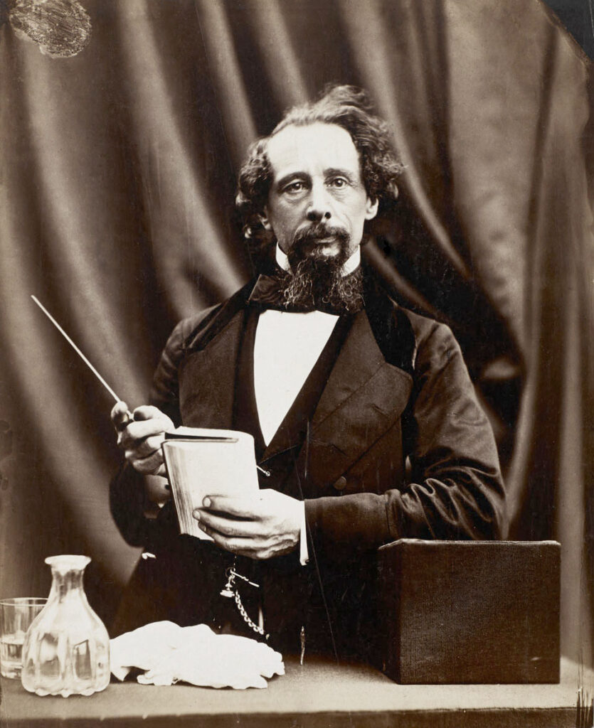 A black and white photograph of Charles Dickens. He is standing at a desk holding a notebook and pen. There is a pair of gloves, a bottle and a glass of water on the desk.