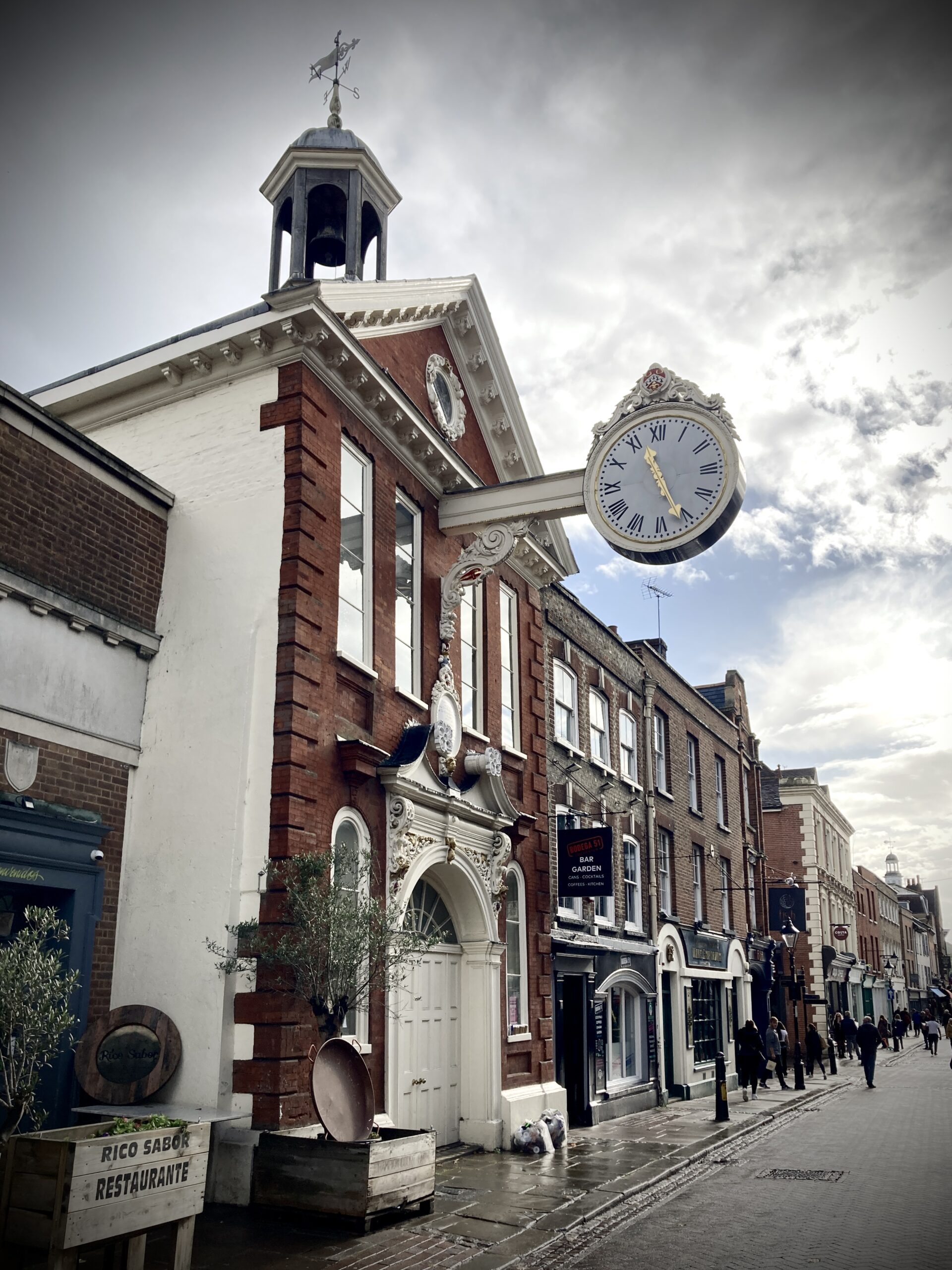 image shows the Old Corn Exchange, Rochester. It is an old red brick building. A large, white, ornate clock projects from the second storey of the building, over the High Street.