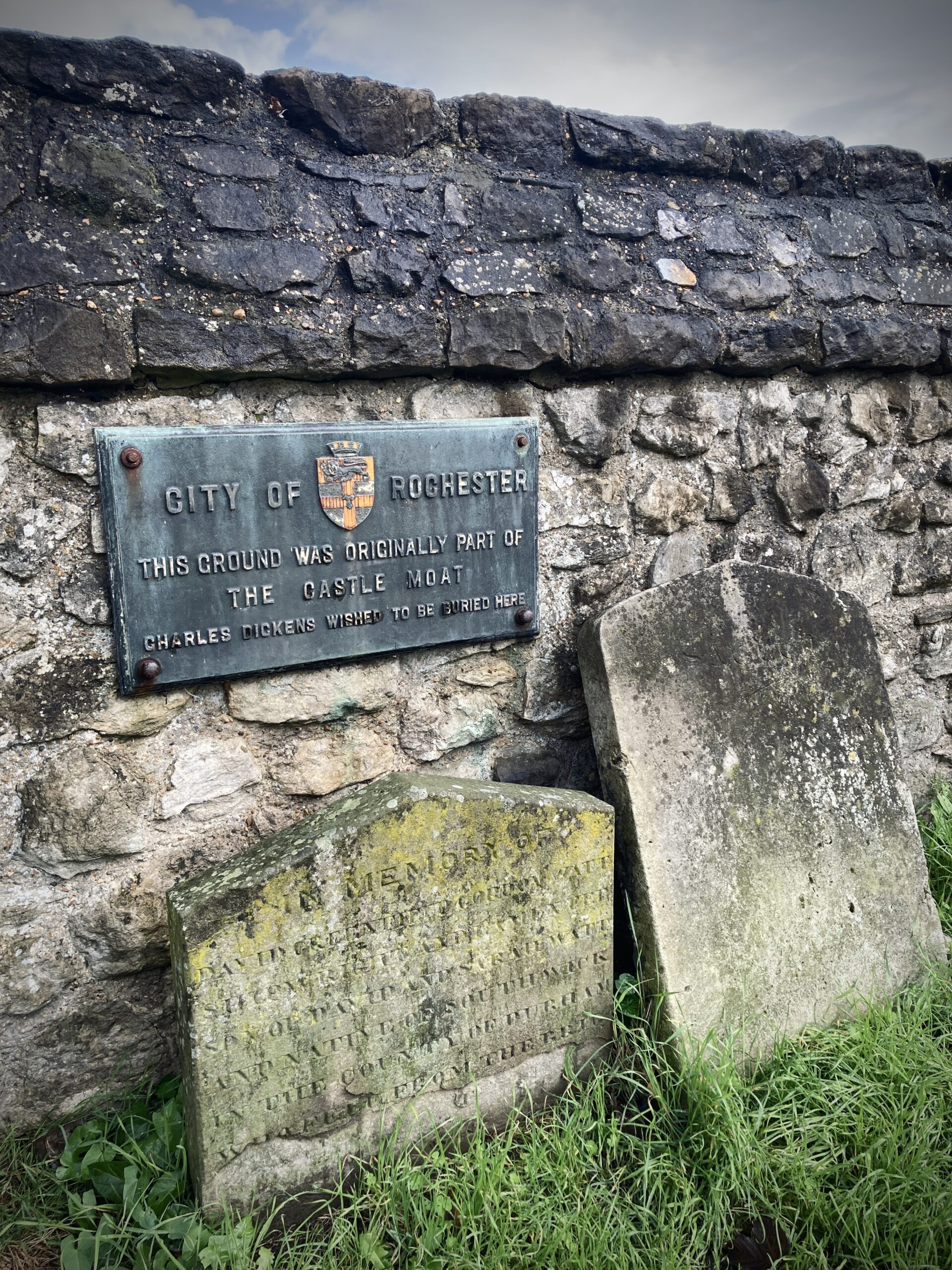 An image showing two weathered gravestones against a wall in the grounds of Rochester Castle. Theere is a plaque on the wall above the graveyards. The words on the plaque are all in capital letters and say, "City of Rochester. This ground was originally part of the castle moat. Charles Dickens wished to be buried here."