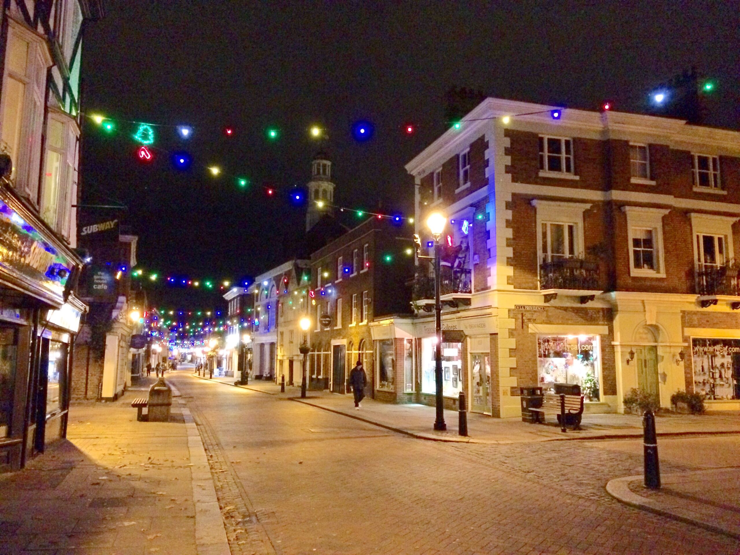 Rochester High Street at night, with Francis Iles Galleries on the right hand, Subway and the Garden House cafe on the left. There are multi-coloured Christmas lights strung between the buildings and a man dressed in black walking down the street.