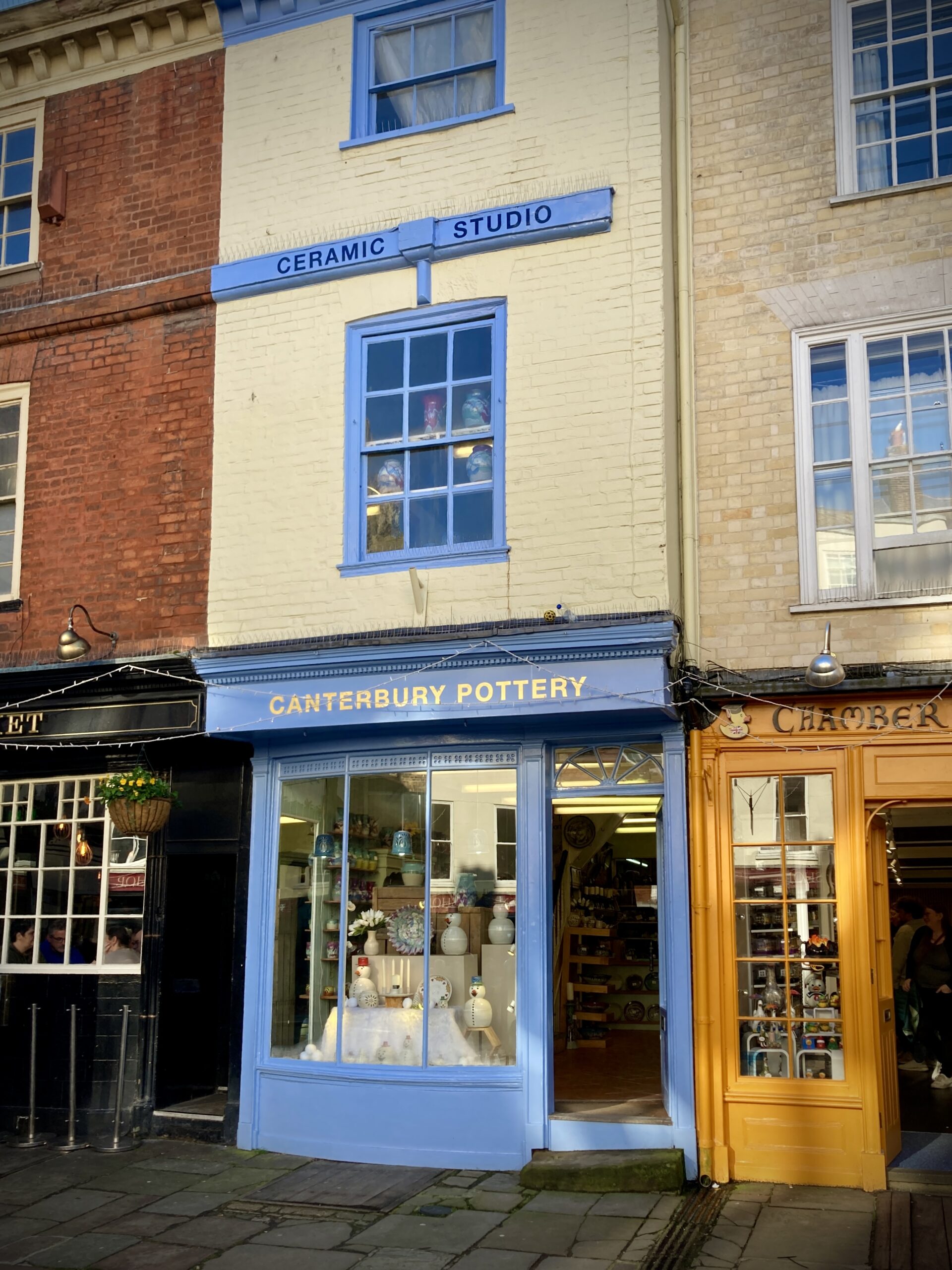 The storefront of Canterbury Pottery. It is a cornflower blue shop with 'Canterbury Pottery' in gold lettering above the window. The window contains ceramic snowman figures, Christmas dishes and other china. On the left side of the shop is a store with a yellow front. On the right is the Old Buttermarket pub.