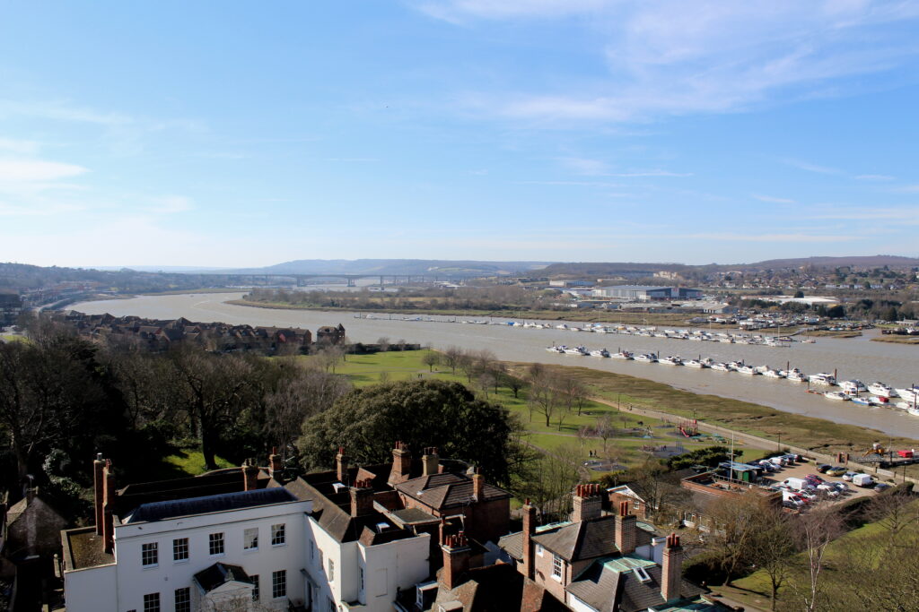 View of the River Medway on a sunny day, taken from Rochester Castle. The river curves away towards the horizon. There are several lines of boats moored on the river. Rooftops of Rochester are in the foreground. The Medway City Estate and Cuxton are visible in the background.