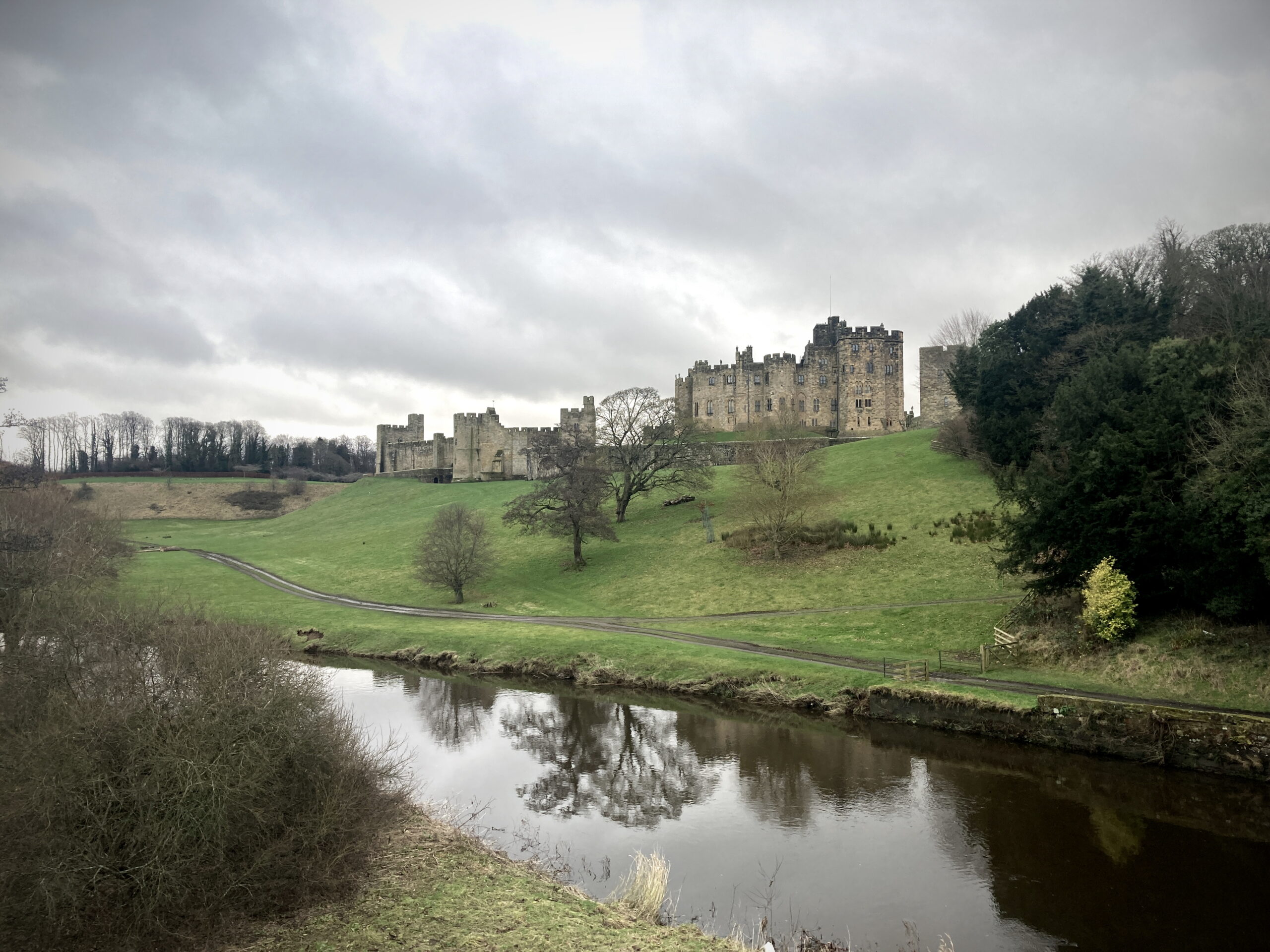 A view of the exterior of Alnwick Castle on a cloudy day. The castle stands on a low grassy hill. The hill is dotted with trees and there is a river at the bottom.