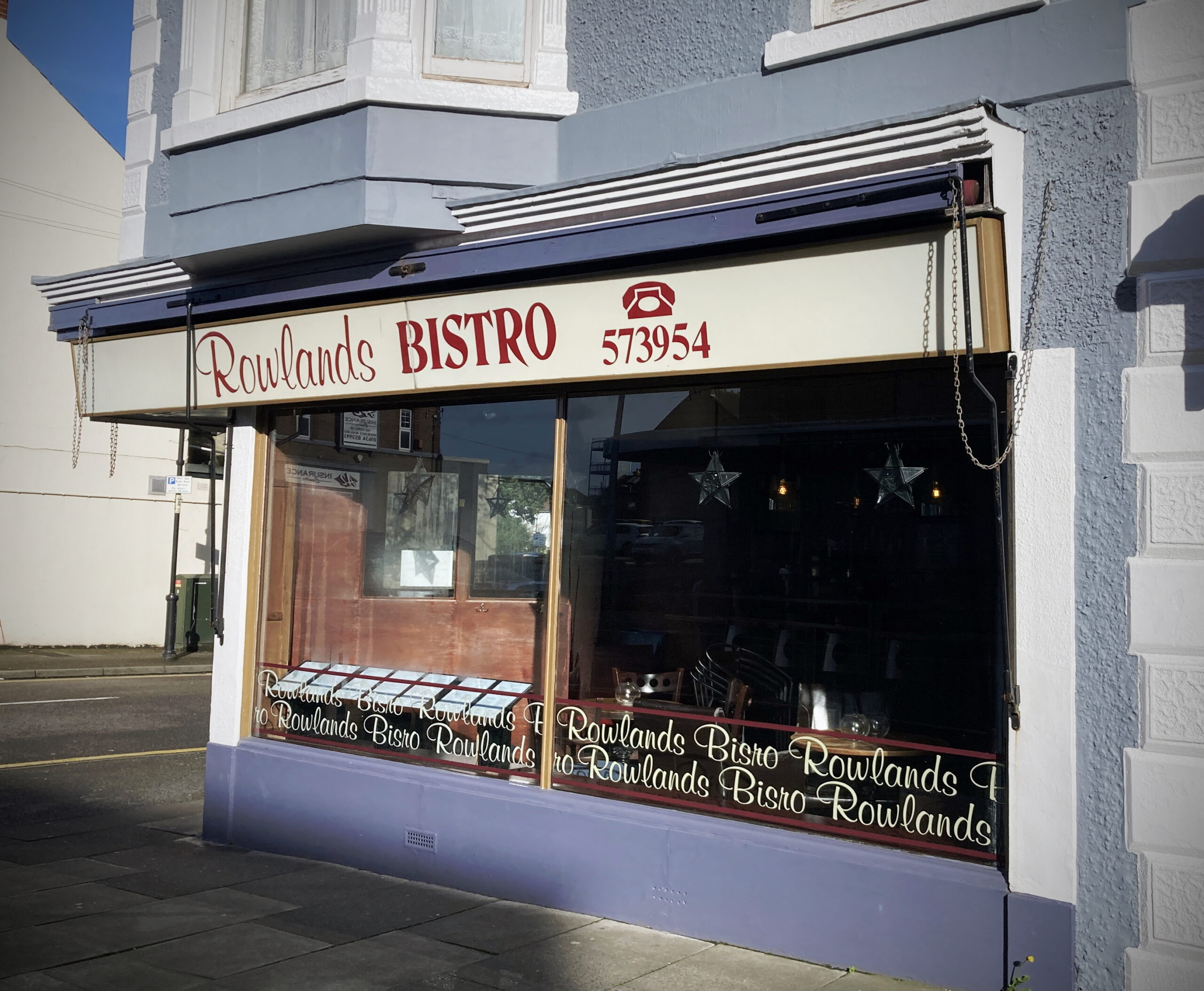 The outside of Rowland's Bistro. It is a pale grey building with large glass windows. The sign above the door says 'Rowland's Bistro' in red text on a pale yellow background. 