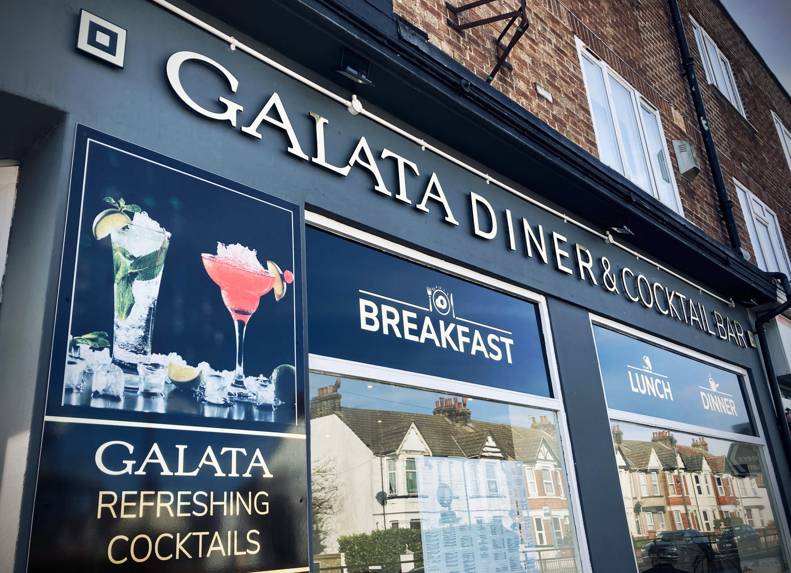 A close up shot of the outside  of Galata. It is a black building with the words 'Galata Diner & Cocktail Bar' in gold capital letters above the windows. Panels above the windows advertise 'Breakfast', 'Lunch' and 'Dinner'. There is a picture of cocktails to the left of the windows.