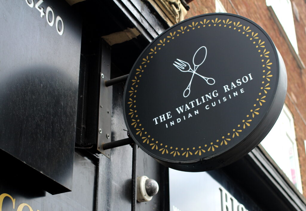 An image of The Watling Rasoi restaurant sign, which reads 'The Watling Rasoi Indian Cuisine'. The sign is black with white text, and a decorative gold border. 