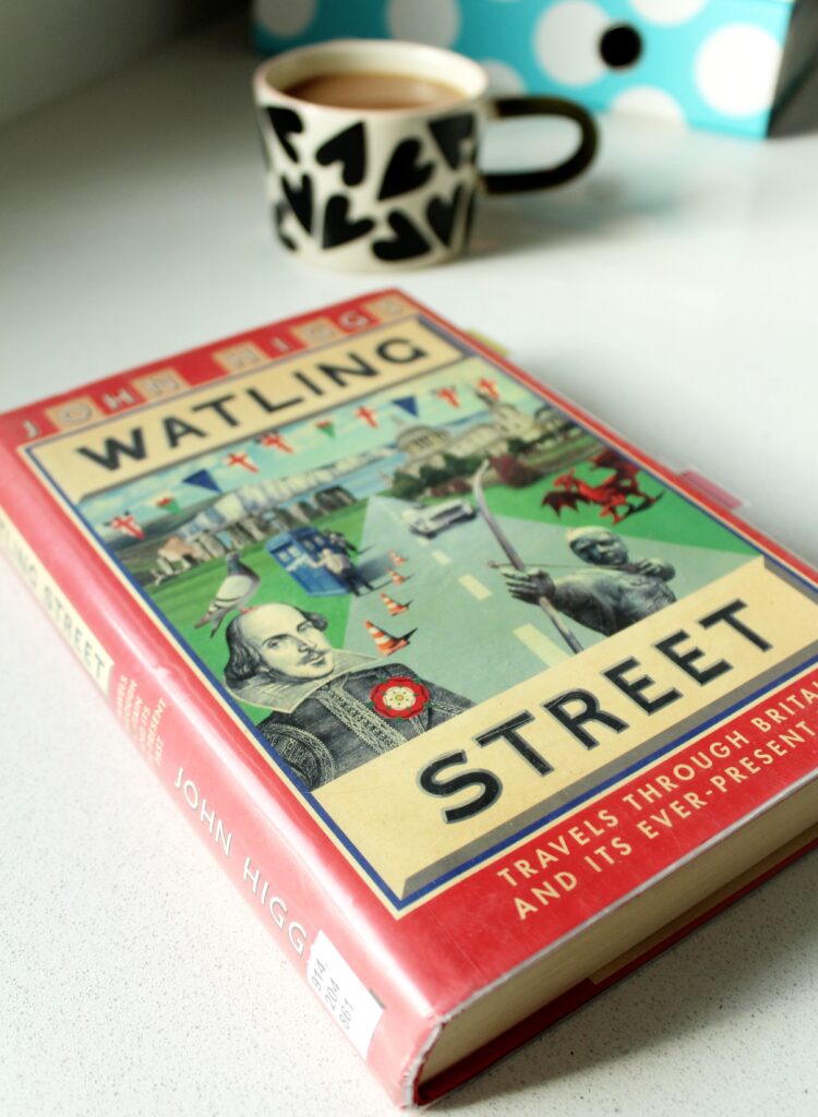 A photo of the book, 'Watling Street - Travels through Britain and It's Ever-Present Past' by John Higgs. The cover is red and illustrated with an image containing pictures of Shakespeare, Robin Hood, traffic cones, a Welsh red dragon and a pigeon. There is a cup of tea in the background of the photo.