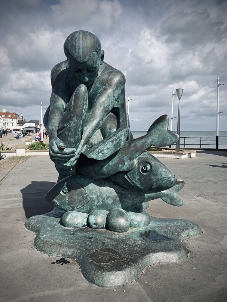 John Buck's bronze sculpture, Embracing the Sea. The sculpture is of a man looking down at a fish he is holding. He is kneeling amid two other fish. The sculpture is green and stands at the entrance to the pier.