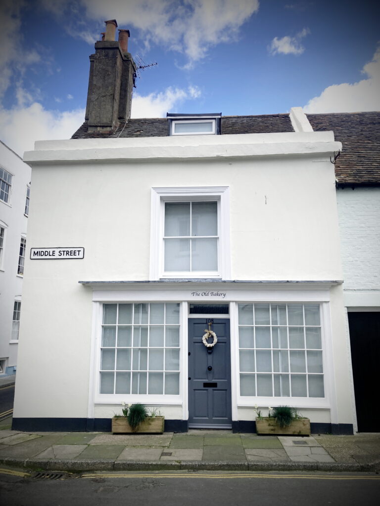 The exterior of The Old Bakery on Middle Street. It is a white, small, two storey Georgian house with a blue door. The words 'The Old Bakery' are above the door. The front door has a wreath on it.  