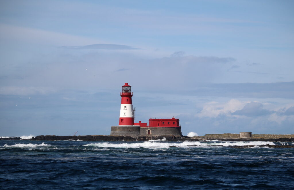 A view of Longstone lighthouse, taken from the boat. The lighthouse is red and white. The sky is blue with light cloud.