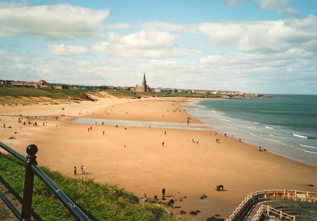 An image of the beach at Tynemouth on a sunny day. The view is across the beach to the church at Cullercoats. The sea is visible on the right hand side and the church centre on the horizon. 