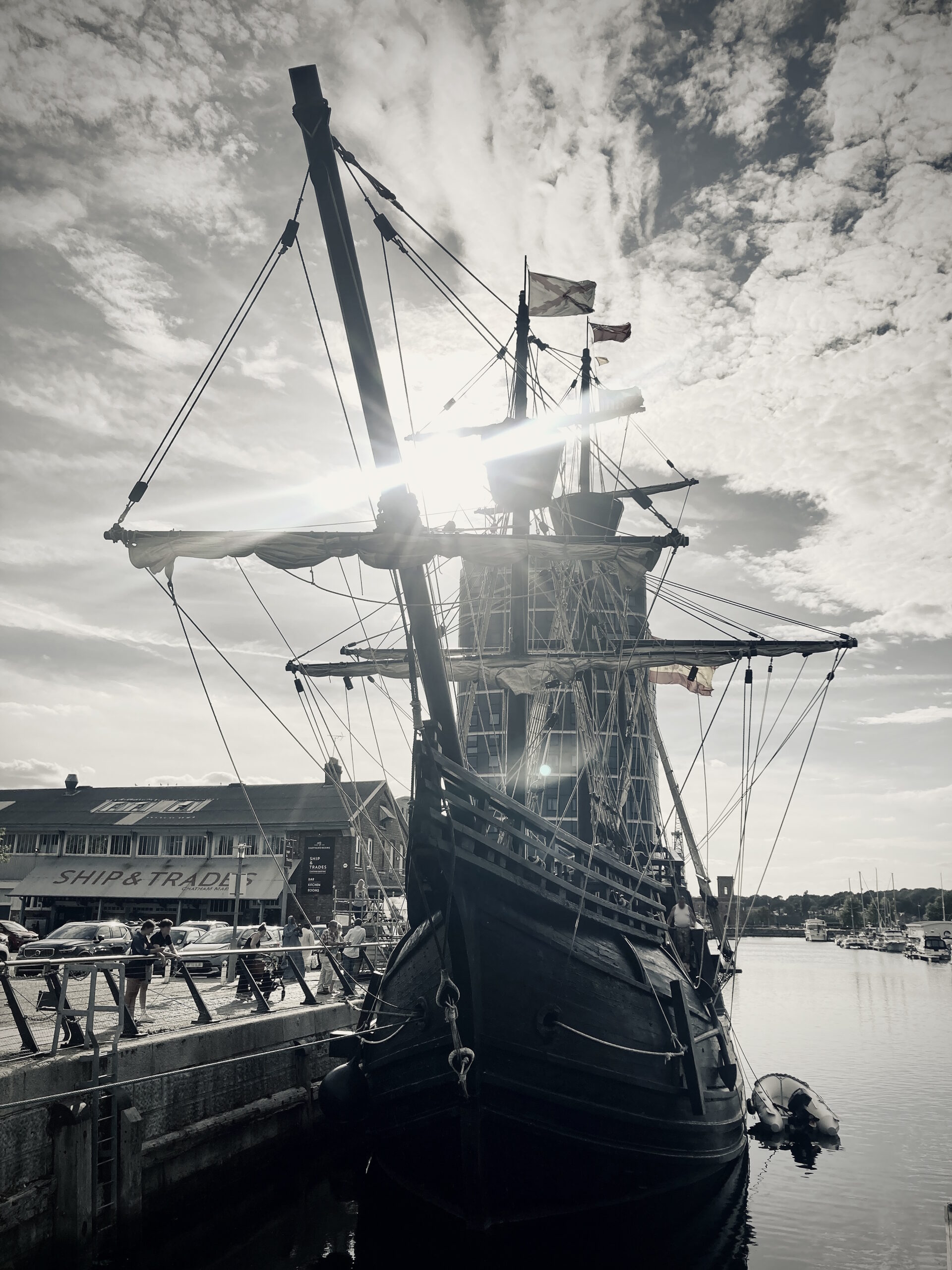 A black and white image of the Nao Victoria moored at Chatham Marina. The Ship & Trades pub in is in the background. People are standing on the dockside looking at the ship.