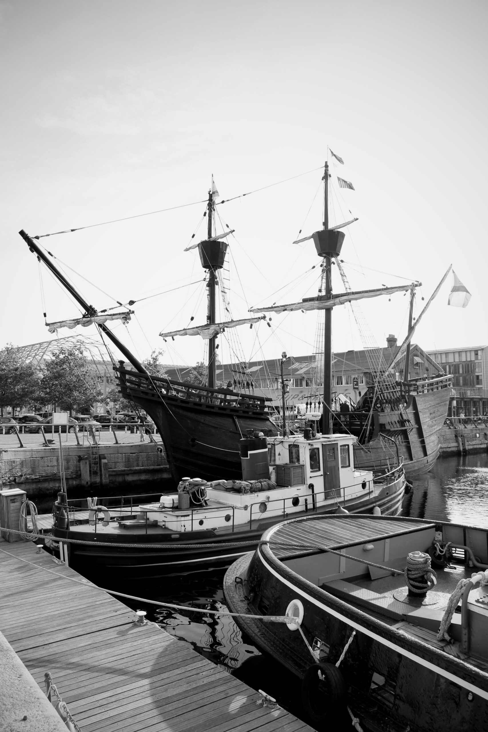 A black and white image of the Nao Victoria at Chatham Marina. Two smaller tugboats are visible in the foreground.