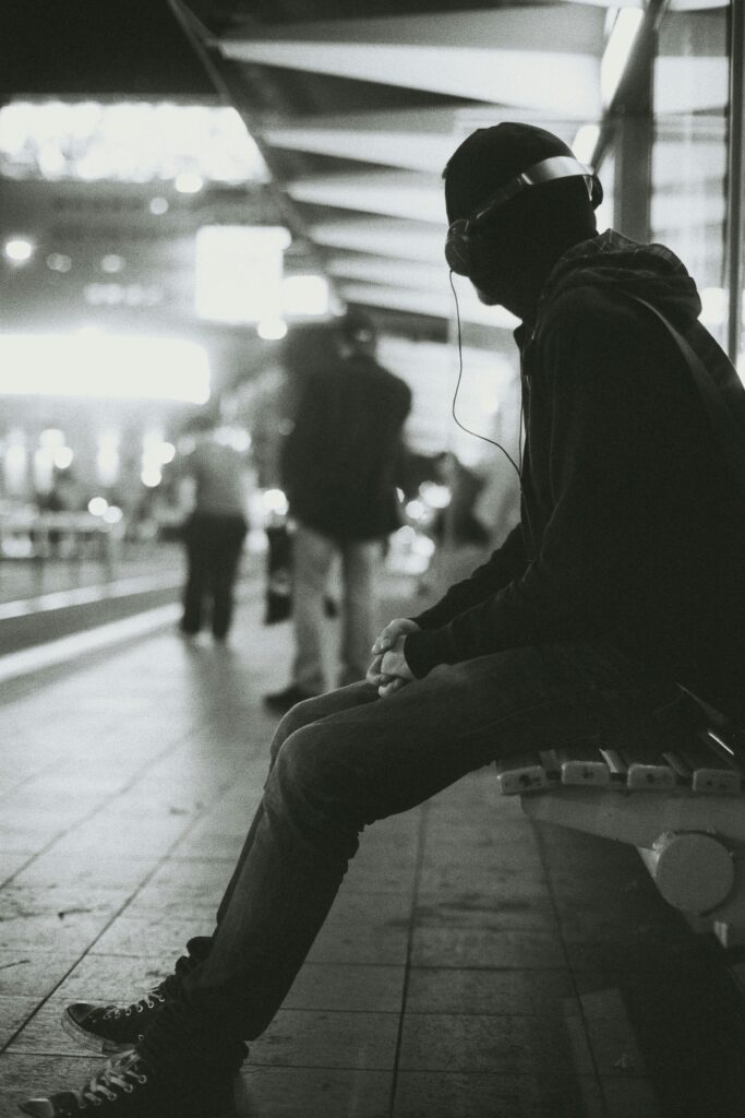 A black and white photo of a man sitting at a station, listening to headphones. The man is wearing a dark hoodie and jeans and is looking away from the camera.