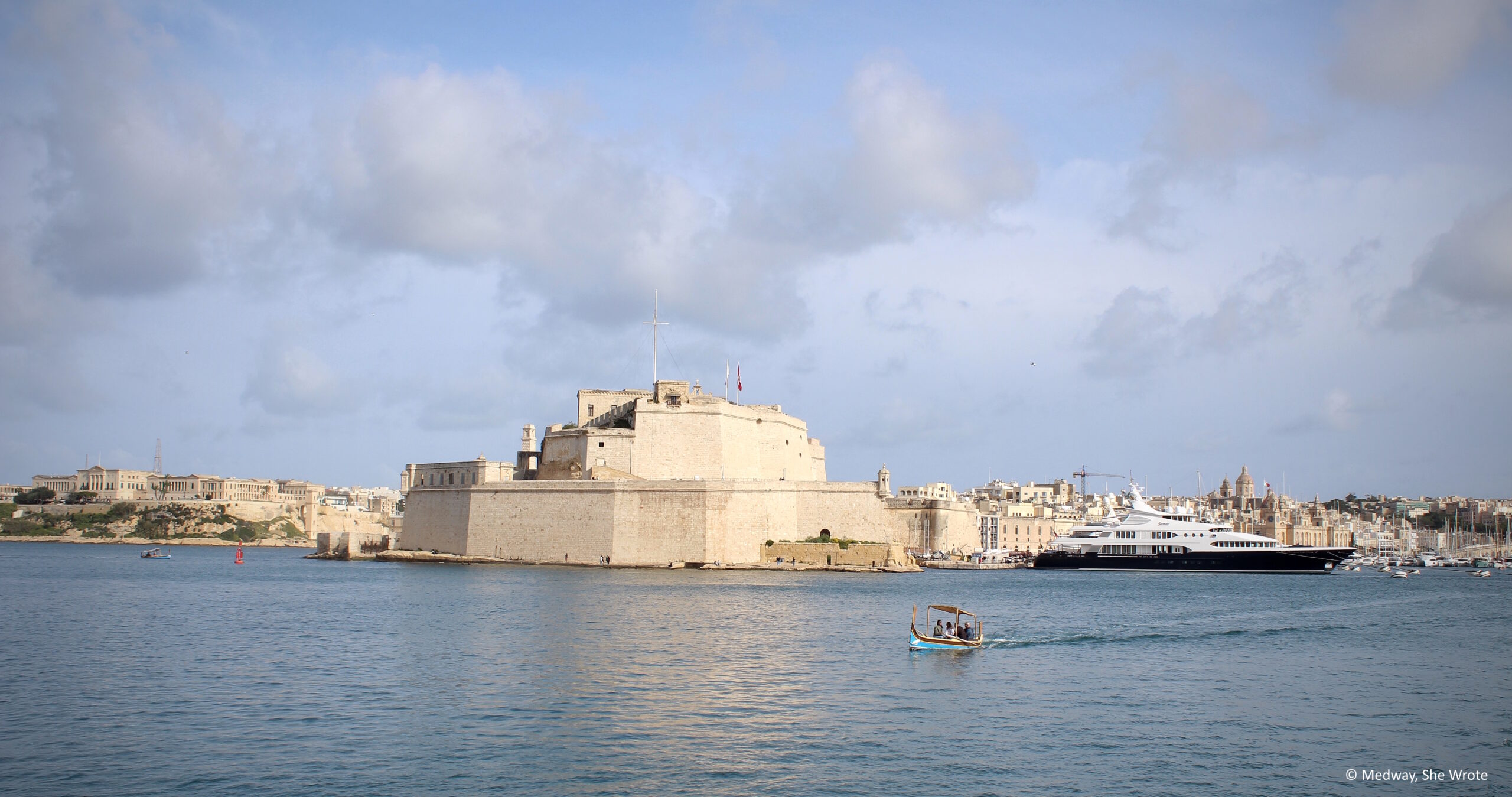 Fort St Angelo viewed from Valletta on a sunny day, with a luzzu boat sailing in the foreground.