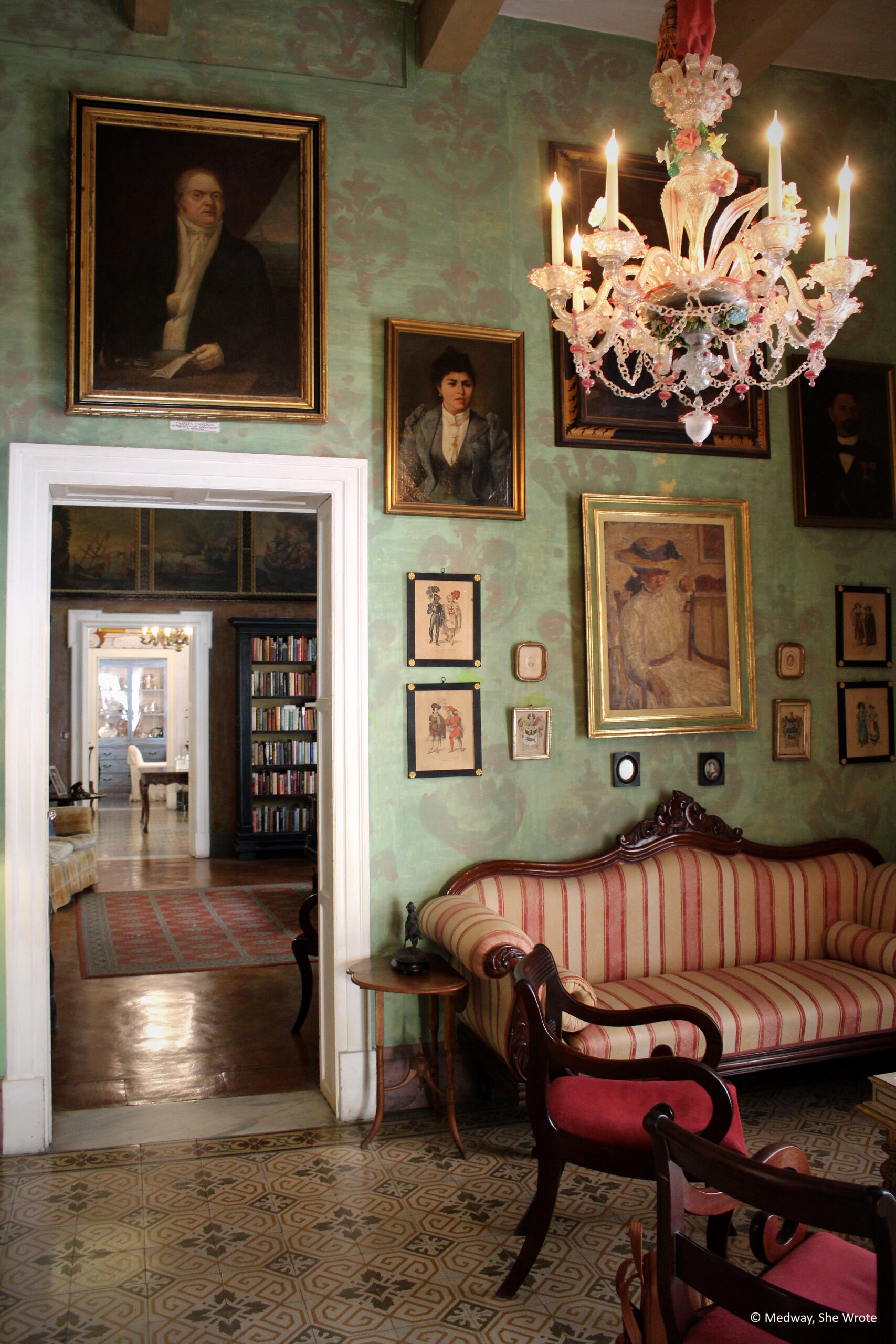 The interior of Casa Rocca Piccola. It is a pale green room with various portraits and artwork on the walls. A red sofa and chairs is standing on the tiled floor. There is a door through which the library next door can be seen.