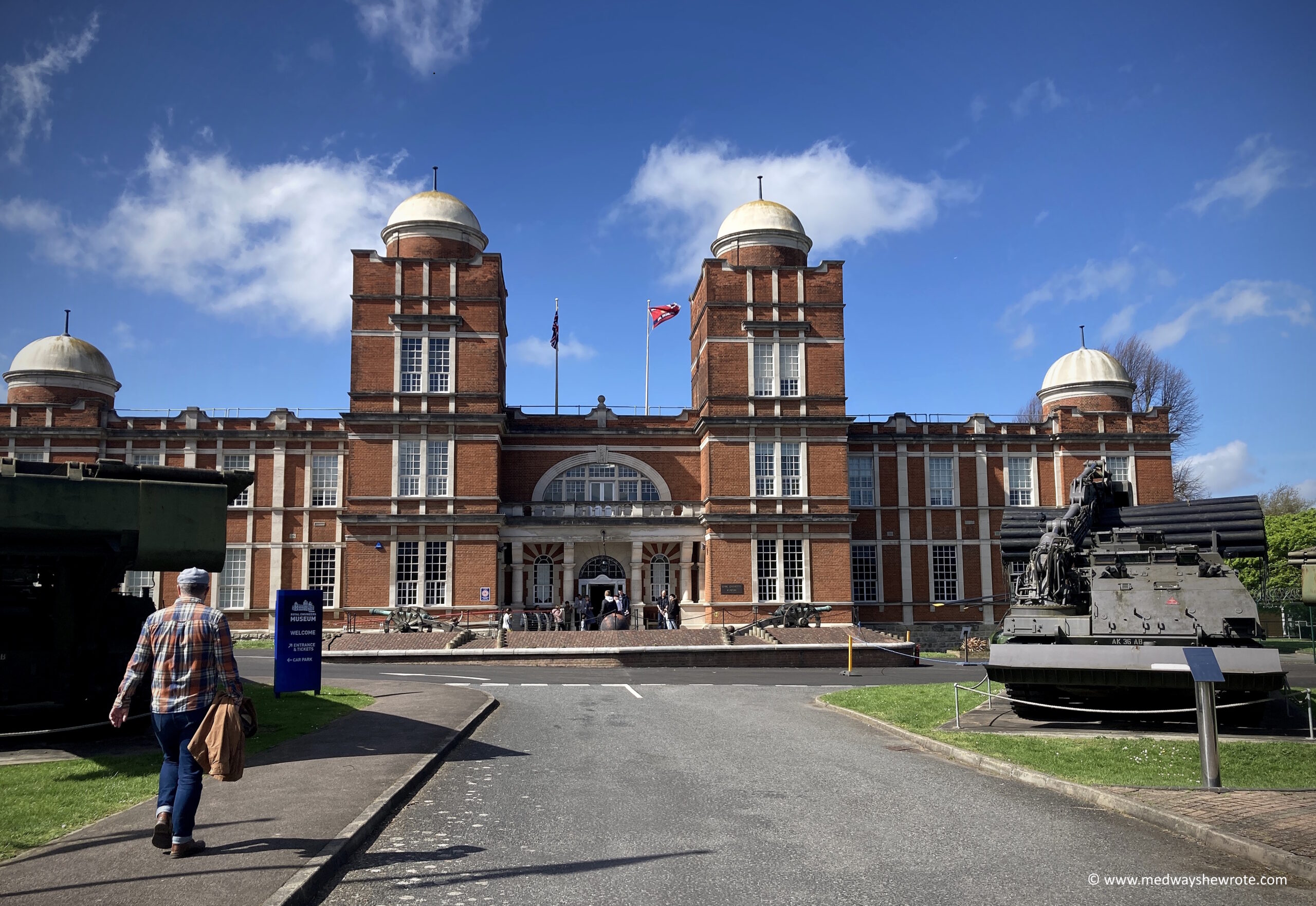 The outside of the Royal Engineers Museum on a sunny day. There are a two tanks and a man walking towards the museum in the foreground.