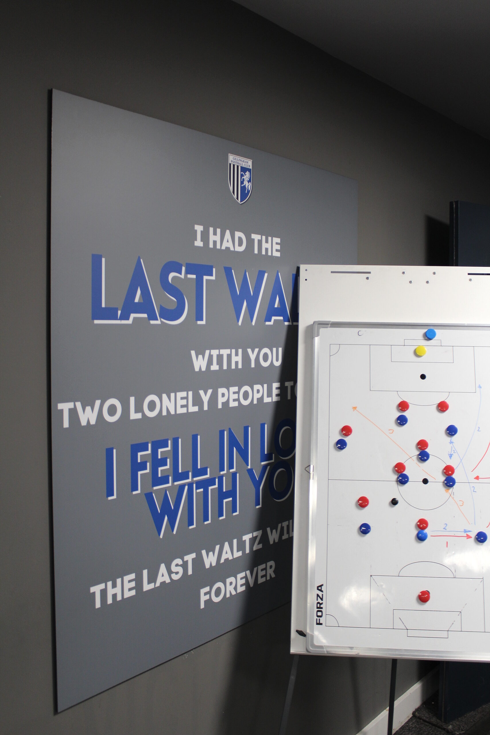 Inside the Priestfield press room. There is a poster on the wall with the text of the Last Waltz set out in blue and white text on a grey background, and a whiteboard with coloured pins marking out positions on a pitch.