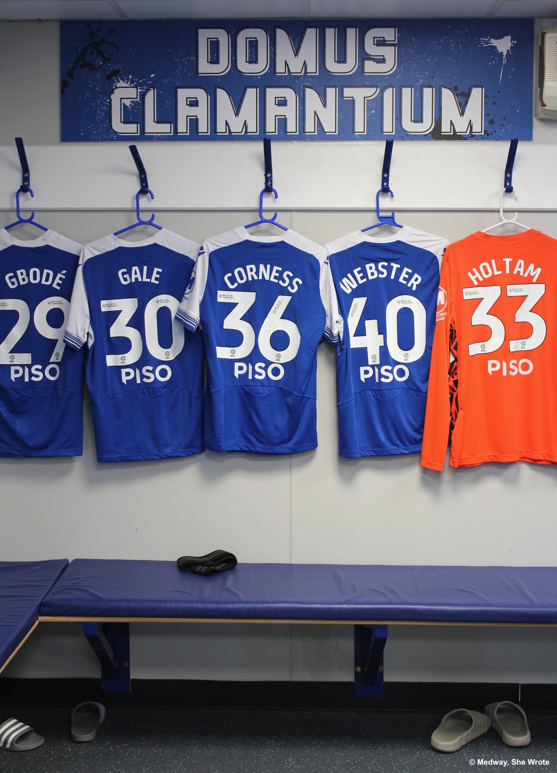 Shirt numbers 29 (Gbode), 30 (Gale), 36 (Corness), 40 (Webster) and 33 (Holtam) hanging up in the players' dressing room. The phrase 'Domus Clamantium' is on the wall above the shirts, in white capital letters on a blue background.