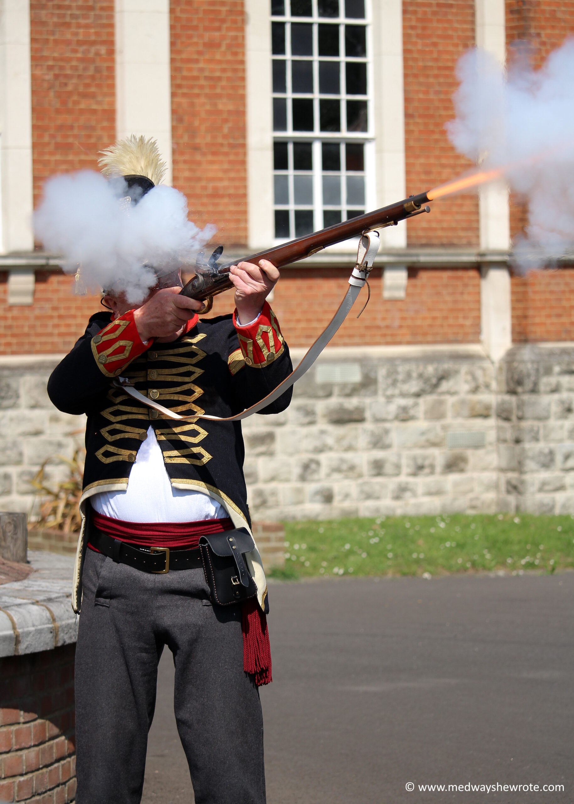 A man dressed as a Napoleonic sapper firing a Flintlock musket in the grounds of the Royal Engineers Museum. The flash of the fired bullet and smoke from the gun are visible. Smoke is obscuring the man’s face.