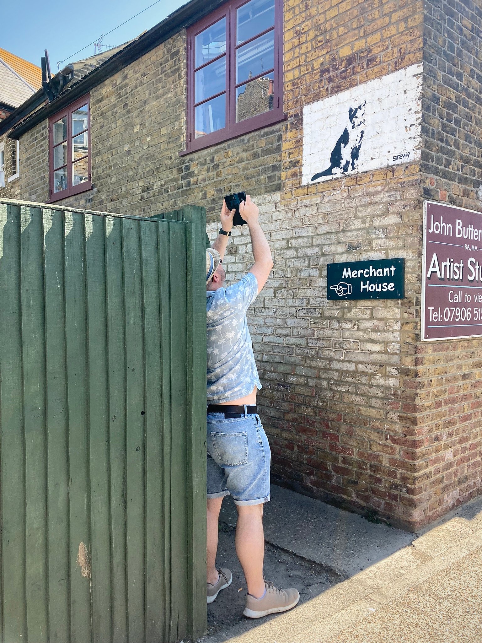 A man taking a photo of a black and white mural of a cat on a wall. He is wearing a straw hat, sage green shirt with palm trees on, pale blue denim shorts and beige trainers. He is standing between a tall green fence and a brick building, holding his phone above his head to take the shot.