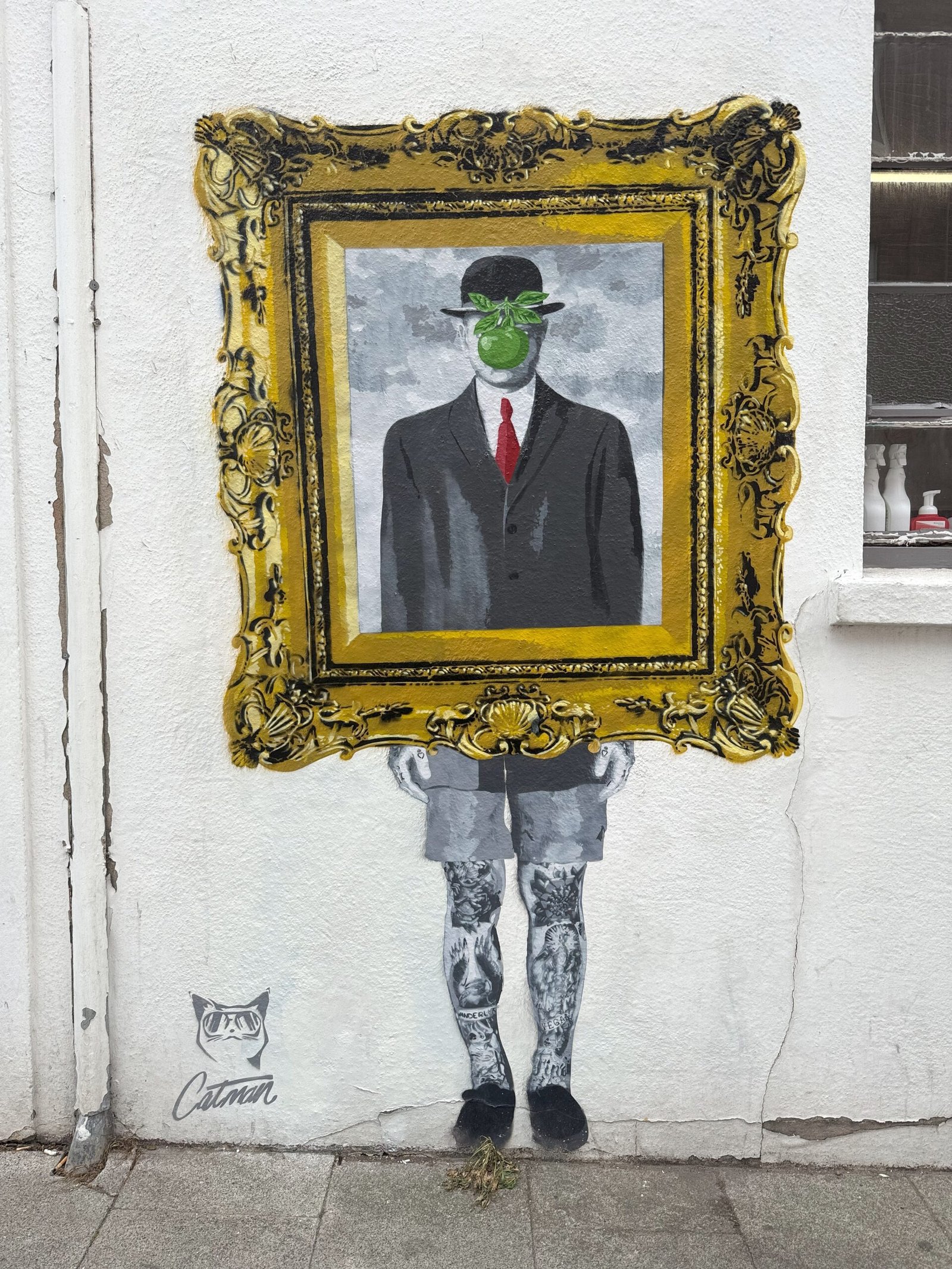 'Son of Inked Man', by Catman. The mural shows a man in a bowler hat and suit inside an ornate gold frame, with a green apple obscuring his face. Below the frame, the man's legs are shown, covered in animal tattoos.