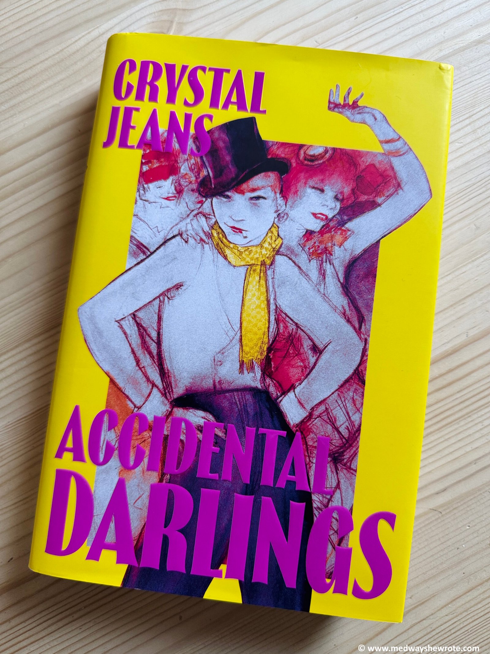 A hardback copy of Accidental Darlings by Crystal Jeans. The cover is bright yellow with dark pink font and depicts a short haired woman wearing a top hat and male 1920s style clothing.