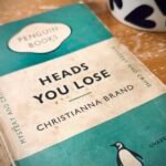 A old, Penguin paperback copy of 'Heads You Lose' by Christianna Brand on a coffee table. The bottom of a mug with a pattern of black hearts on a white background is visible in the background.