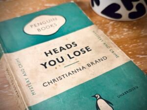 A old, Penguin paperback copy of 'Heads You Lose' by Christianna Brand on a coffee table. The bottom of a mug with a pattern of black hearts on a white background is visible in the background.