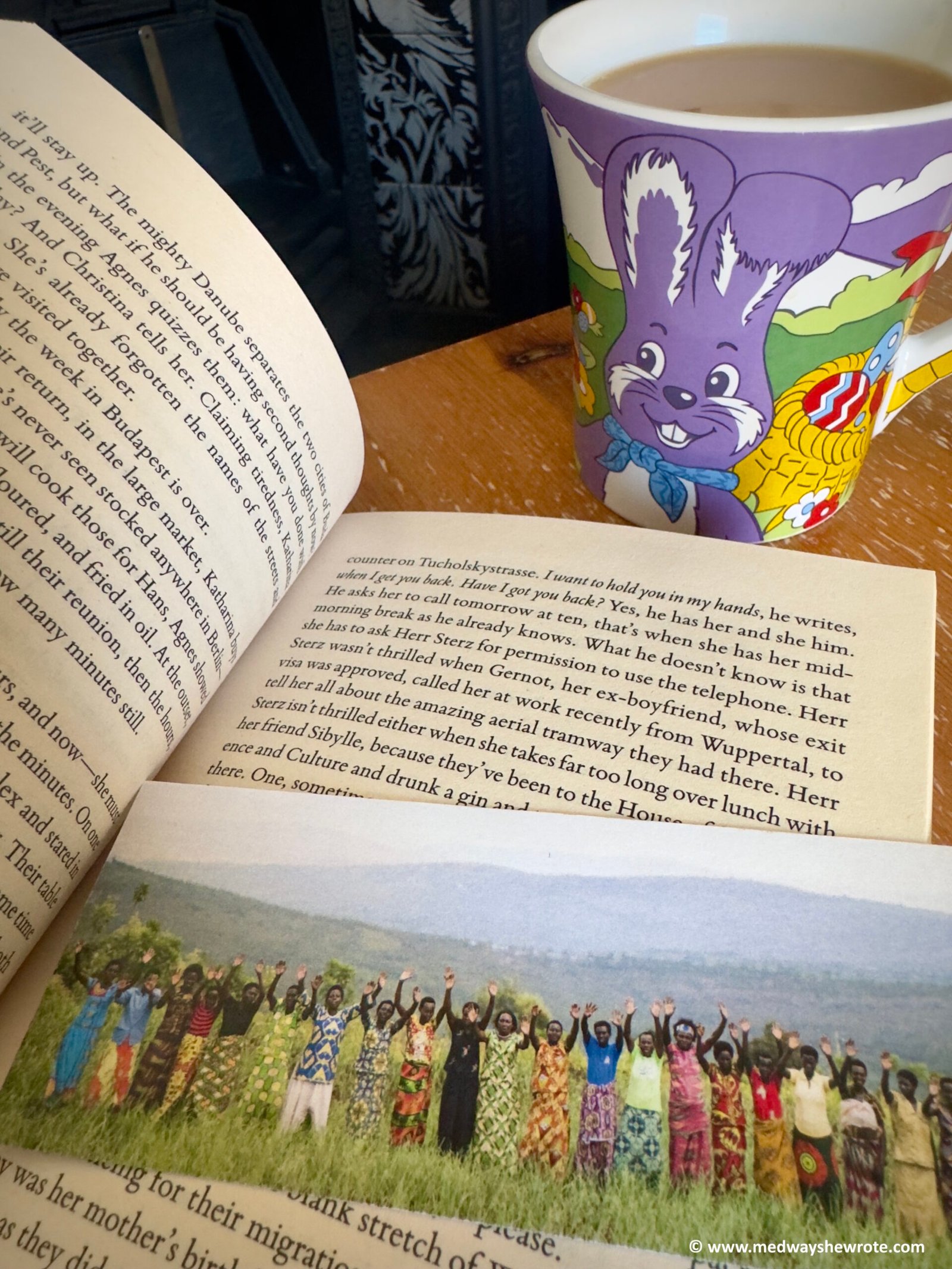 A paperback copy of Kairos by Jenny Erpenbeck, open at page 39. A bookmark showing women in Africa lies across the page. A mug of tea featuring a purple Easter bunny is in the background.