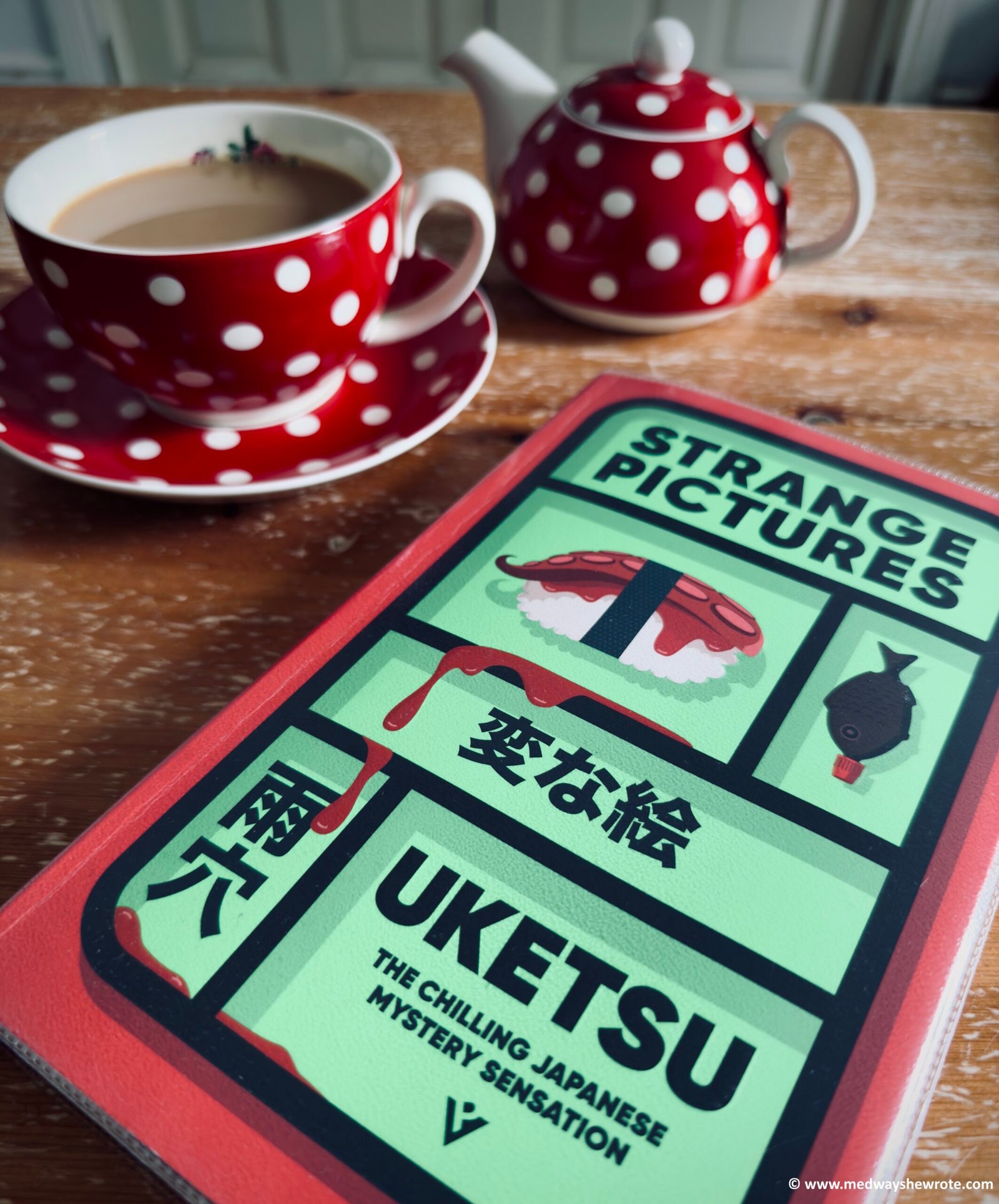 A paperback copy of Strange Pictures by Uketsu, on a coffee table. A small red teapot with white spots, and a matching teacup and saucer, is in the background.