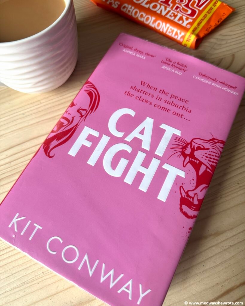 A hardback copy of Cat Fight by Kit Conway on a table. A Chocolonely chocolate bar and cup of tea are partly visible in the background. The book has a pink cover with white font, and depicts a big cat and a woman screaming.