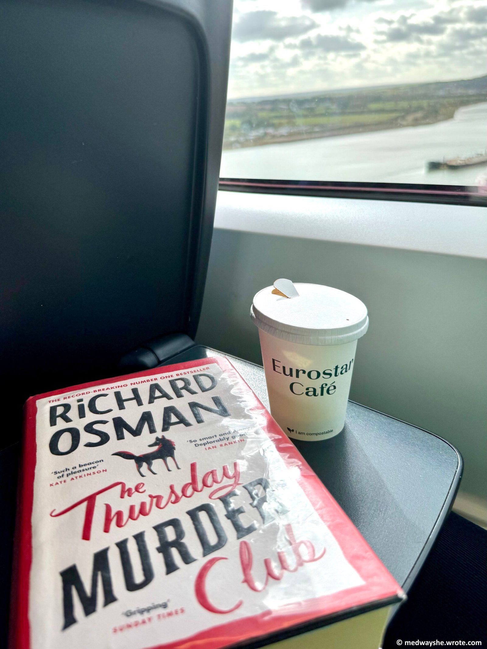A hardback copy of The Thursday Murder Club by Richard Osman on a Eurostar train table. The river Medway is visible from the train window and there is a takeaway coffee cup with 'Eurostar Cafe' on it on the table next to the book.