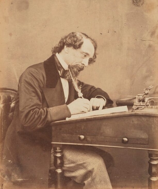 A sepia photograph of Charles Dickens writing with a quill pen at a desk.