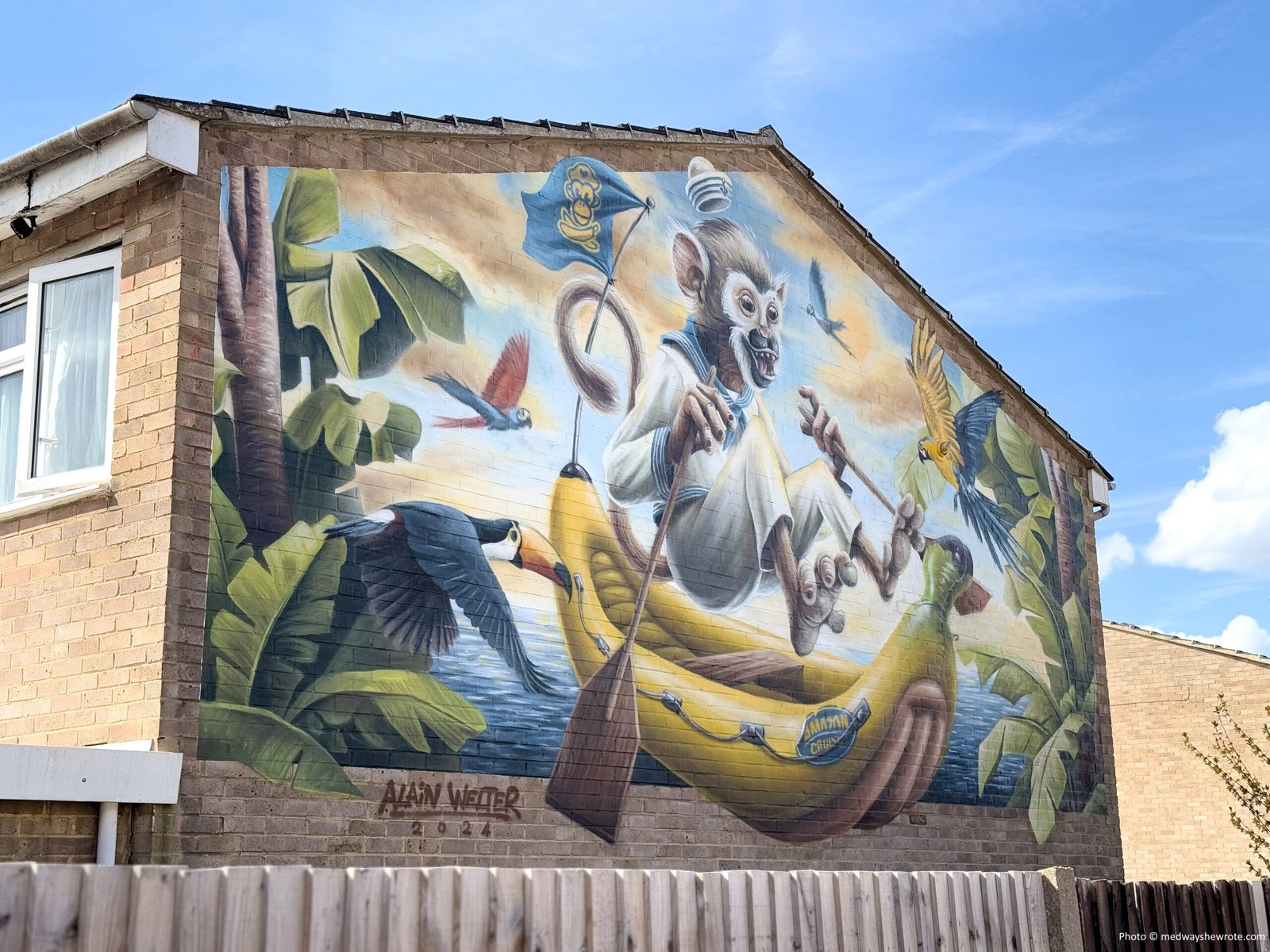Alain's Welter's Pet Shop mural on the side wall of a house. The mural shows a Humboldt monkey rowing a boat shaped like an inflatable banana, surrounded by tropical birds and palm tree leaves.