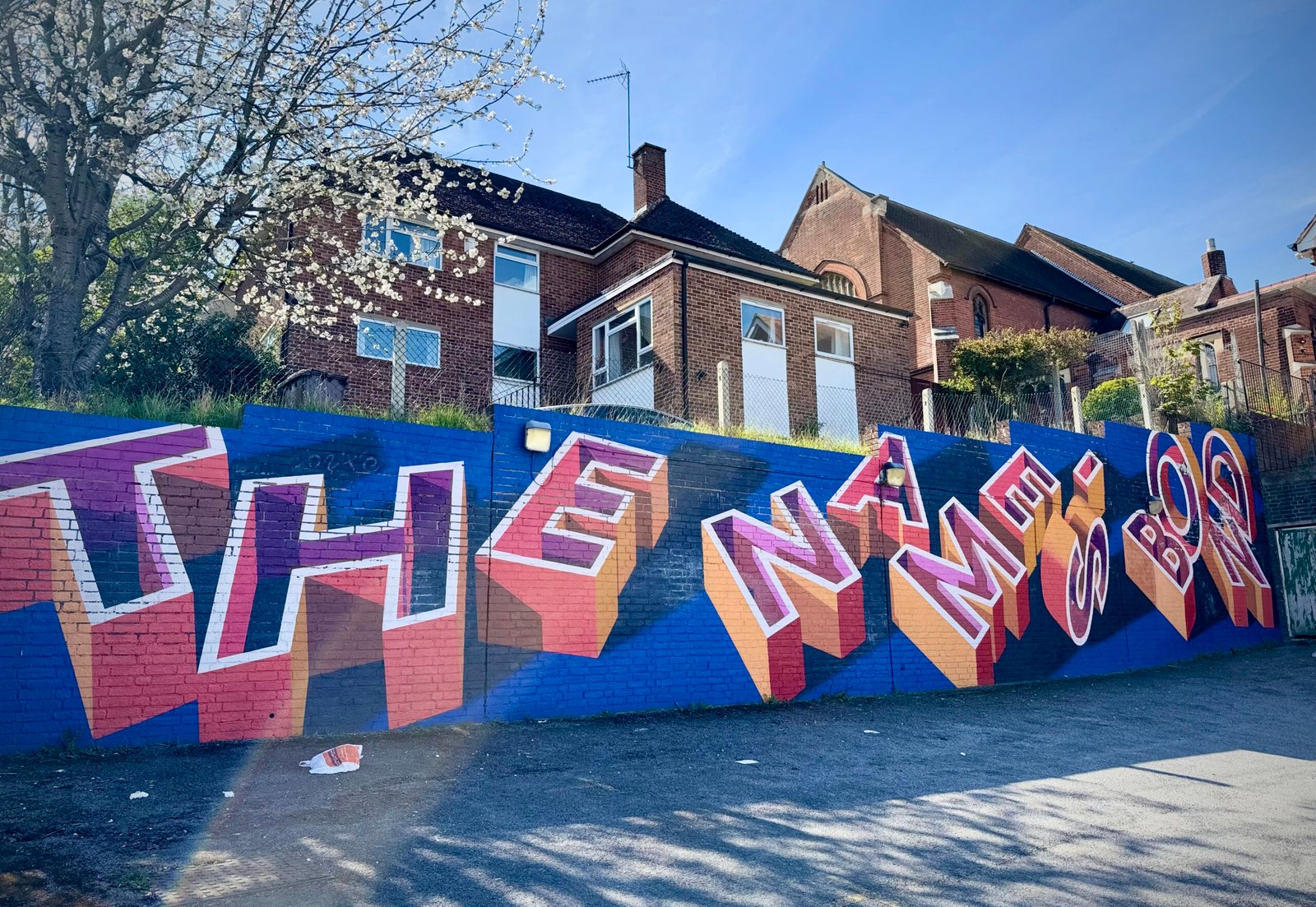 Luke Smile's mural tribute to George Bond. The words 'The Name's Bond' are painted in block capitals in orange, red and purple with white outlines, on a dark blue background on a low wall.