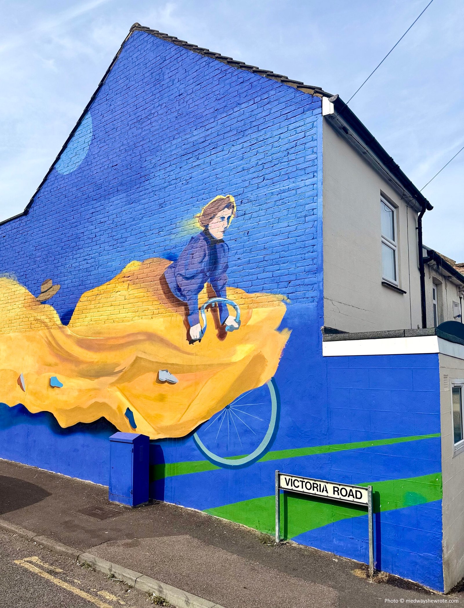 Laura Richardson mural by Hixxy. It shows Richardson on a bike, her huge yellow skirt billowing behind her. Shoes and her hat have blown away behind her. The background is bright blue. 