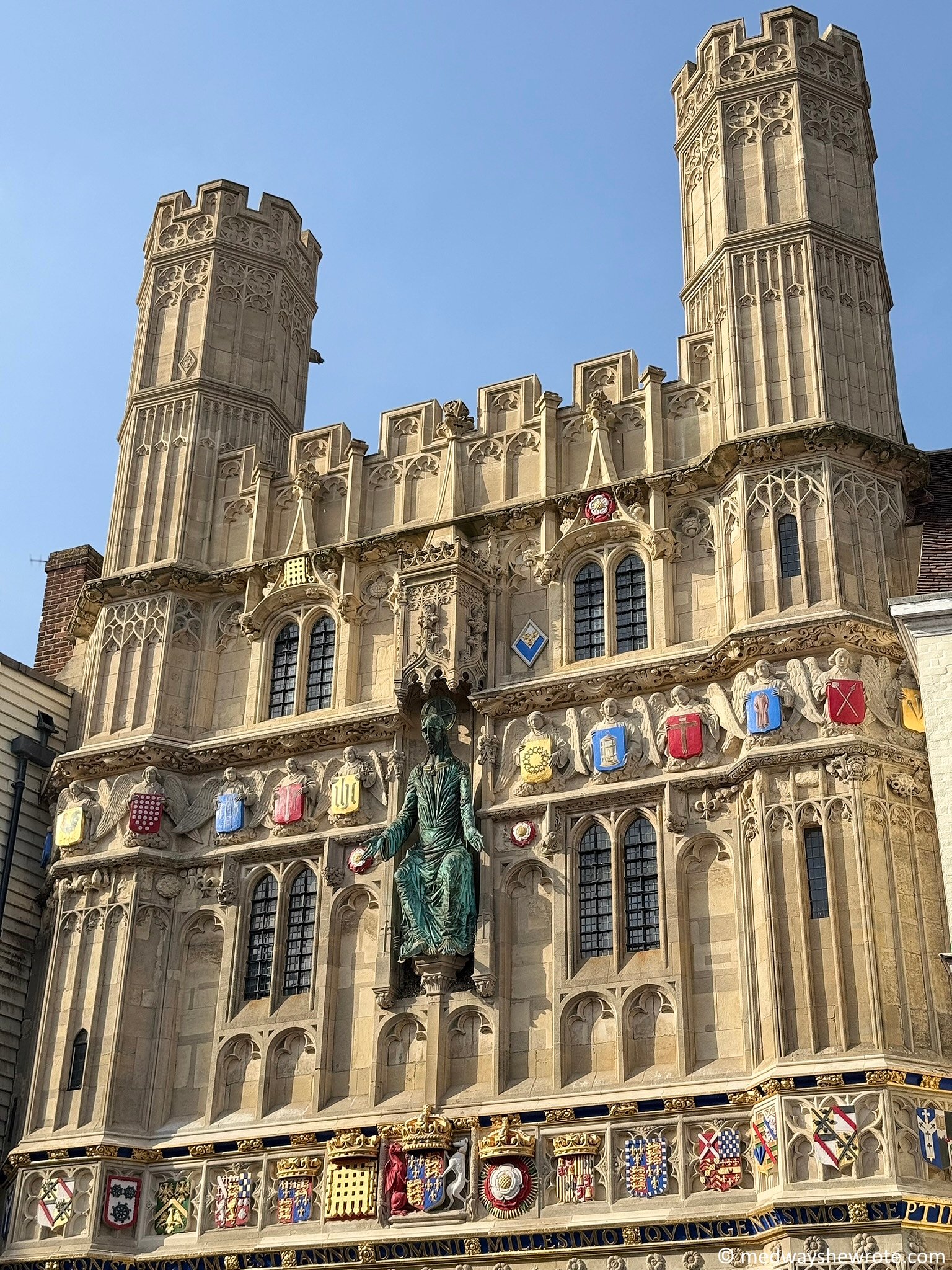 The entrance gate to Canterbury Cathedral. It is constructed of pale yellow stone and decorated with a large bronze sculpture of Jesus and angels holding coloured shields.