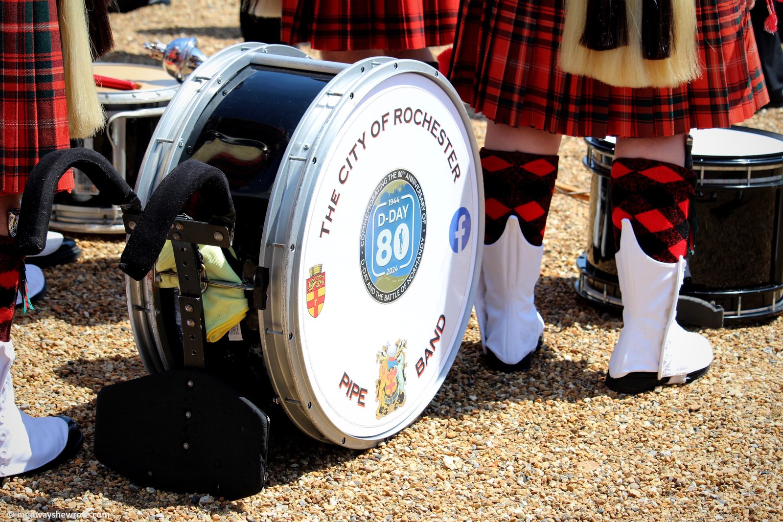 A City of Rochester Pipe Band drum on the parade ground. The white boots of two members of the band, and the bottom of their red tartan kilts, are visible standing next to the drum.