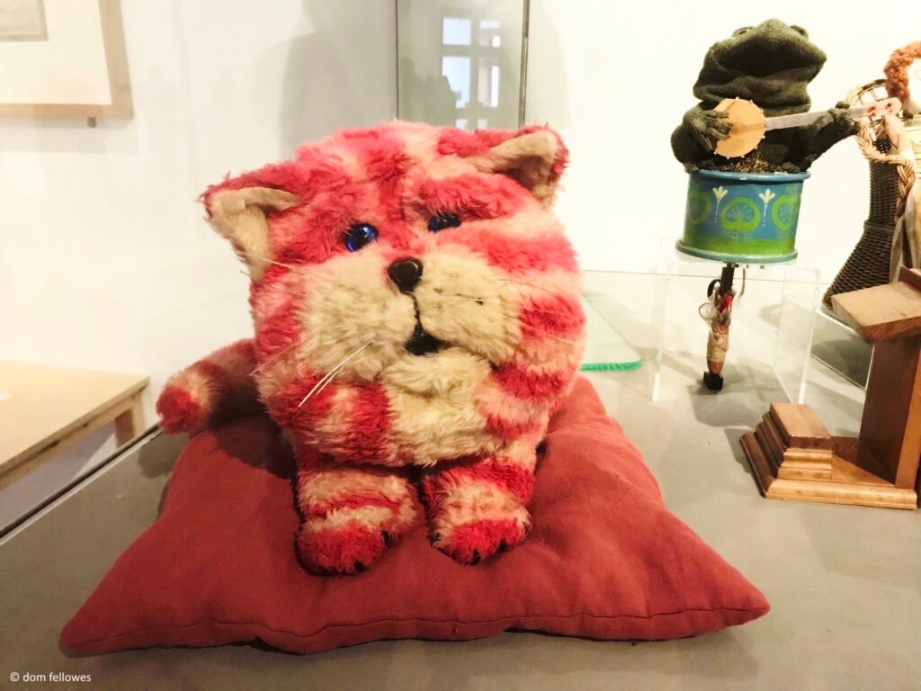 Bagpuss, a pink and white striped cat puppet, on display. He is lying on a red cushion inside a glass display case.