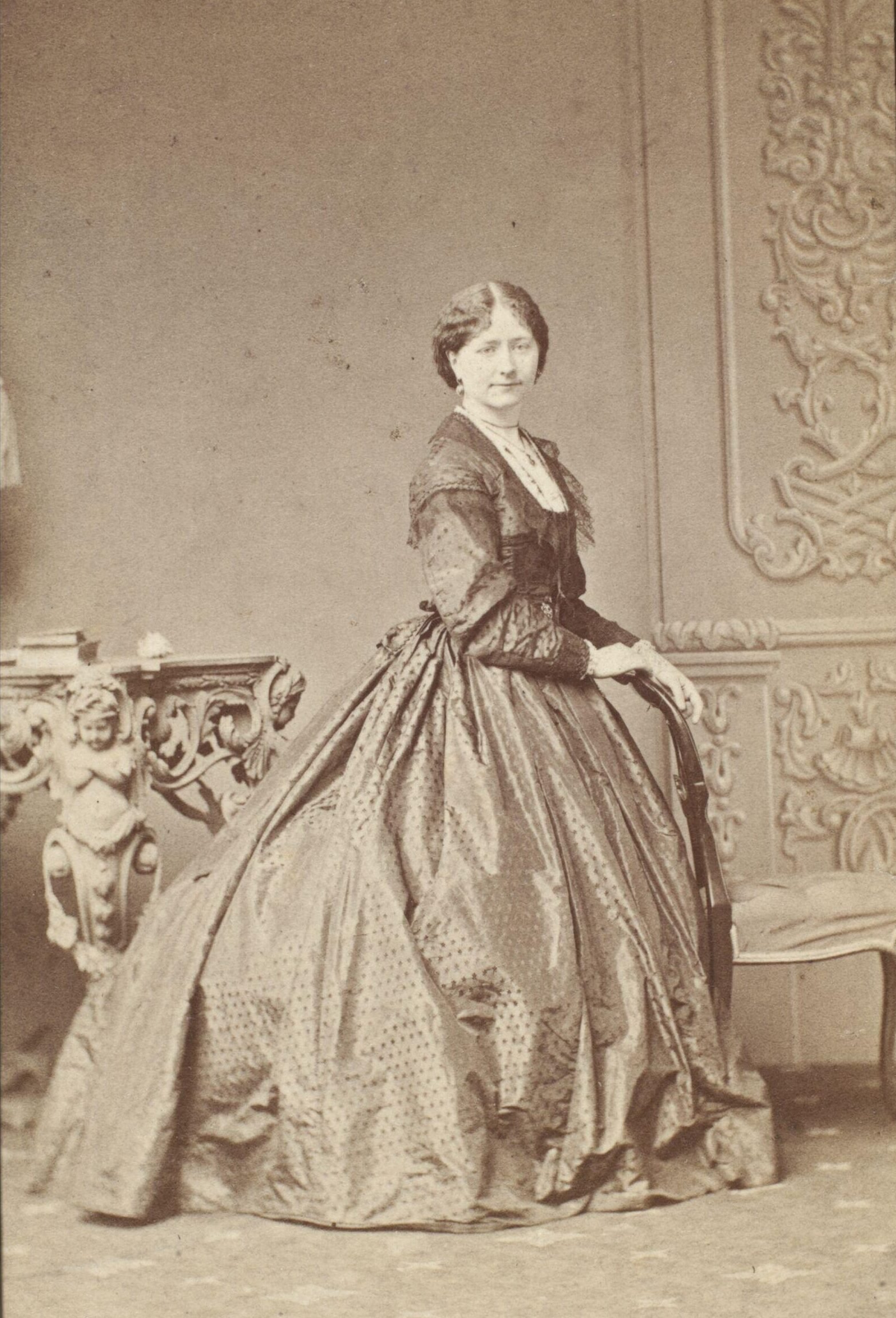 A sepia photograph of Nelly Ternan. She is standing facing the camera, wearing a long gown. 