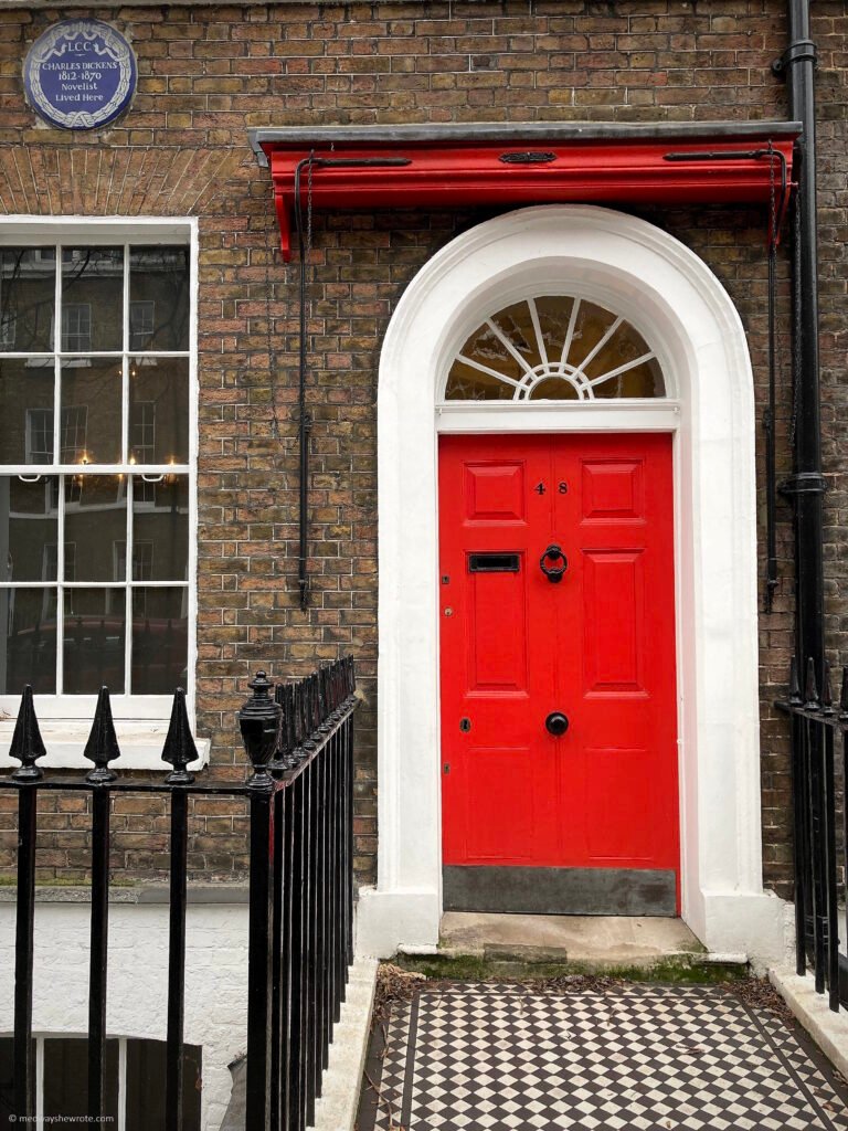 Extertior of 48 Doughty Street. The house has a bright red front door, black railings, and a black and white tiled path leading to the door. There is a blue plaque on the wall that reads, "LCC Charles Dickens, 1812-1870 Novelist Lived Here."