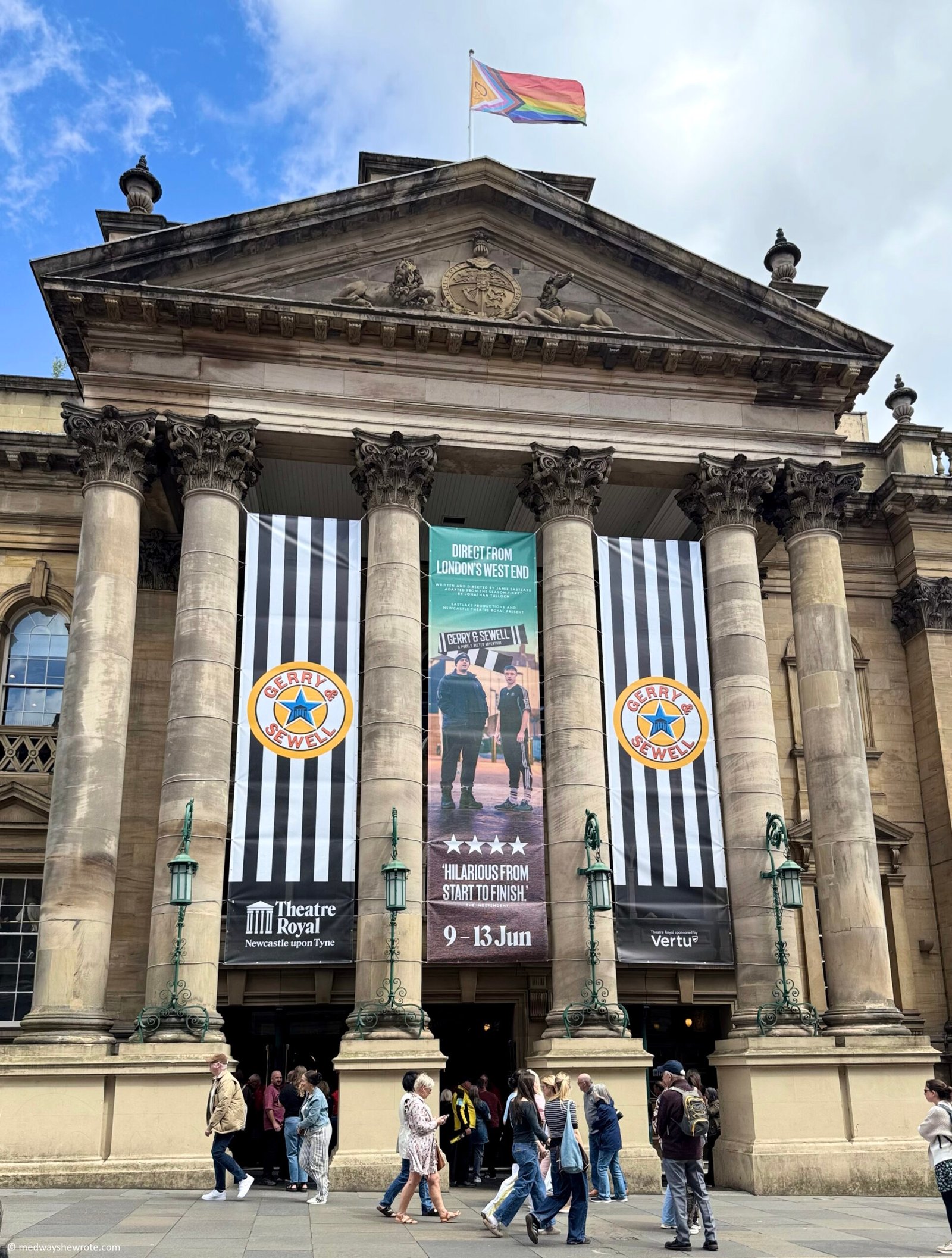 Exterior of Newcastle's Theatre Royal, a Georgian building with a large, pillared entrance. Black and white banners advertising Gerry & Sewell hang between the pillars. It is a sunny day and people are walking on the pavement in front of the theatre. The theatre is flying the Pride flag from its roof.