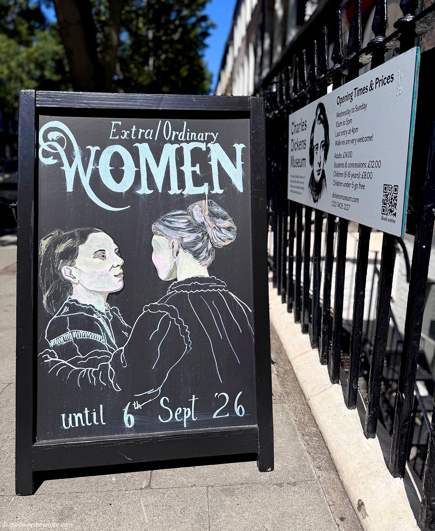 A sandwich board advertising the Extra/Ordinary Women exhibition on the pavement outside the Charles Dickens Museum. The board includes a chalk drawing of Katey and Mamie Dickens turning towards each other, with text 'Extra/Ordinary Women until 6th Sept 26'.