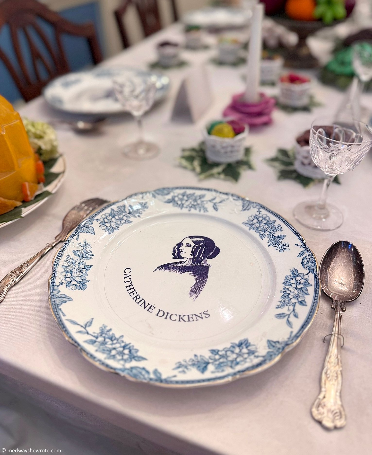 A white china plate printed in blue with an image of Catherine Dickens. The plate has a blue floral border and the words 'CATHERINE DICKENS' printed on it. The plate is on a laid table in the dining room of the Charles Dickens Museum.