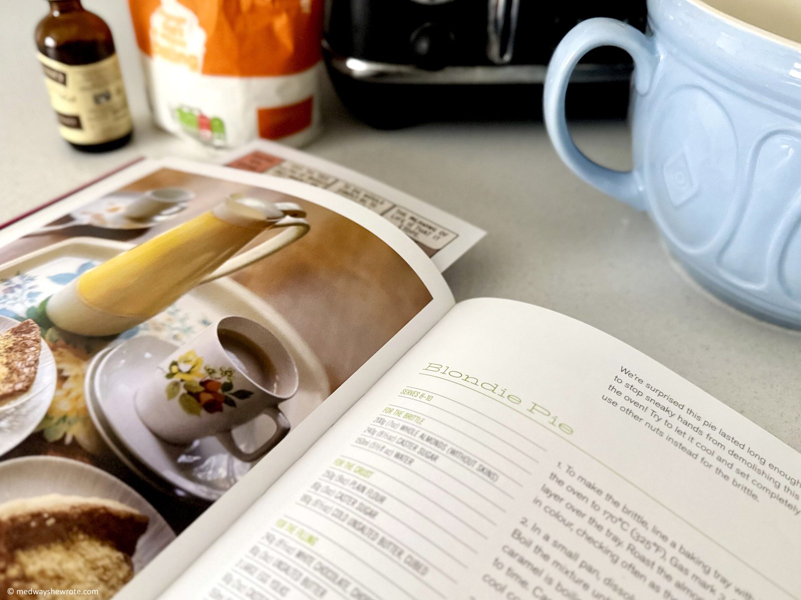 An open recipe book on a kitchen counter. The book is open at a recipe for Blondie Pie. A pale blue mixing bowl, baking ingredients and part of a toaster are visible in the background.